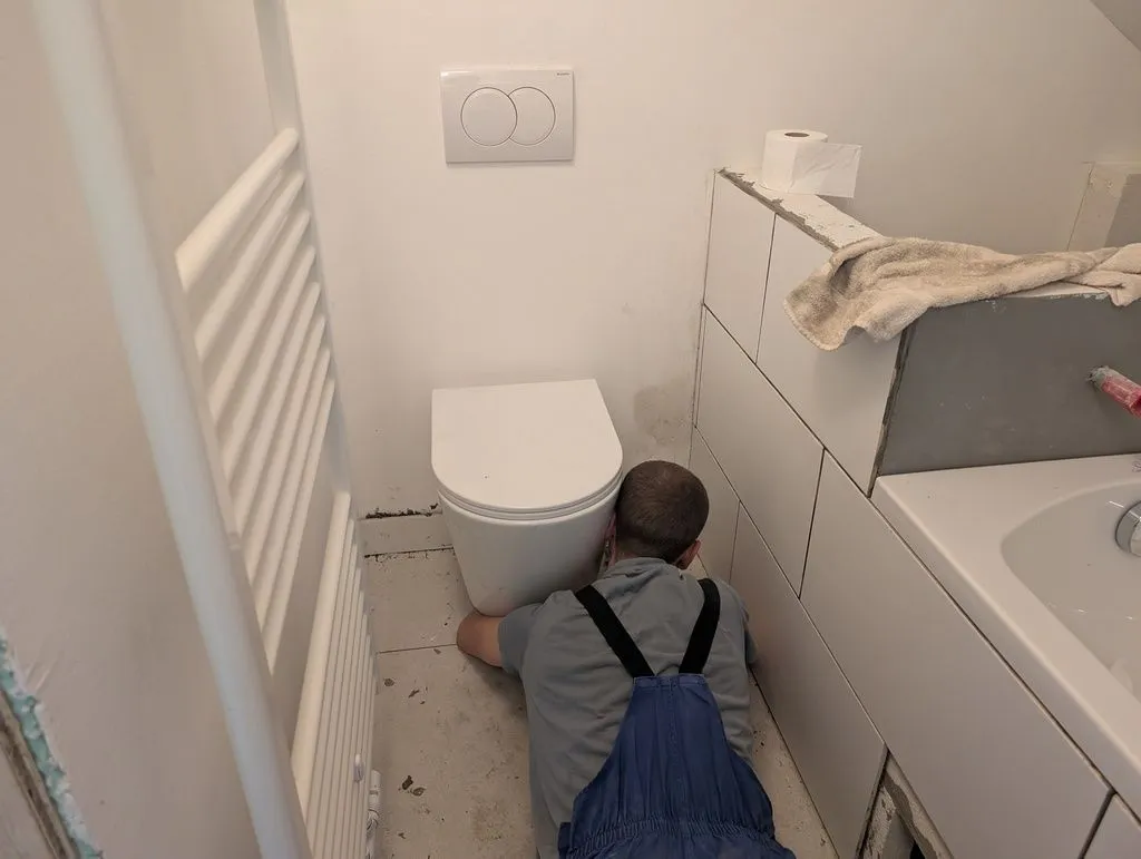 Applying skirting/baseboard tile around the perimeter of the bathroom floor, including under the toilet.