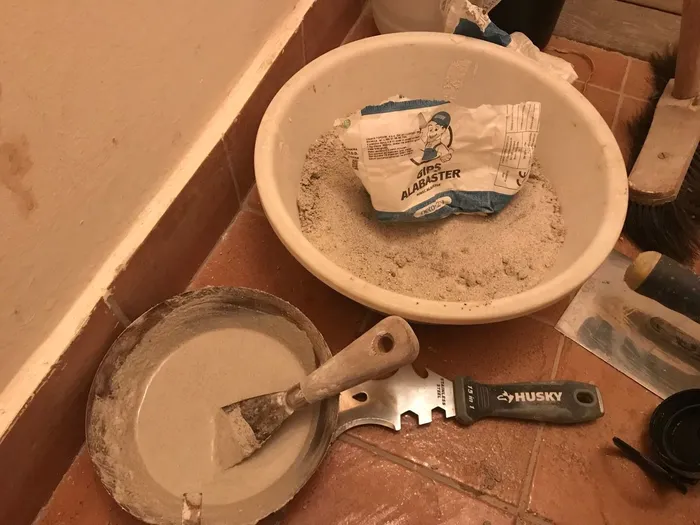 Plastering materials layed out on the floor: spackle knife, plaster, and bucket for mixing.